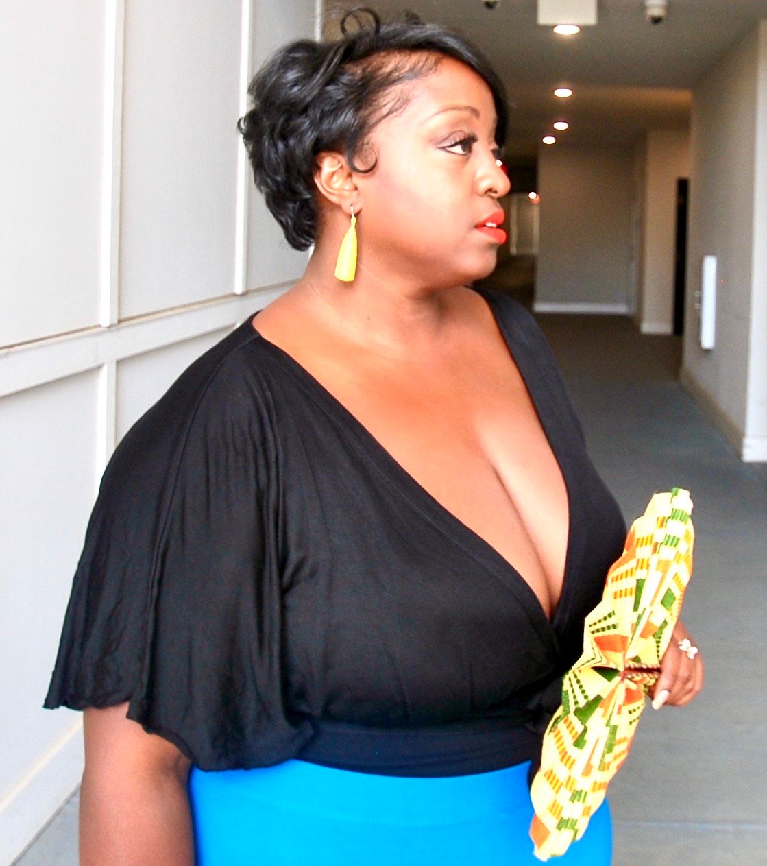 Image of Dominique Bercy. She's dressed in a black top and blue skirt holding a kente cloth-inspired fan, looking away from the camera.